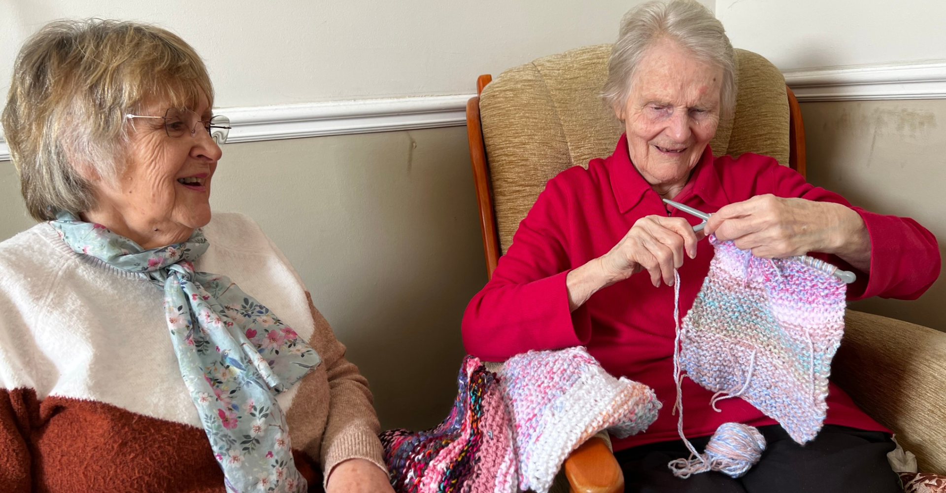 Kathy (befriender) sits next to Margaret, who is knitting a scarf.
