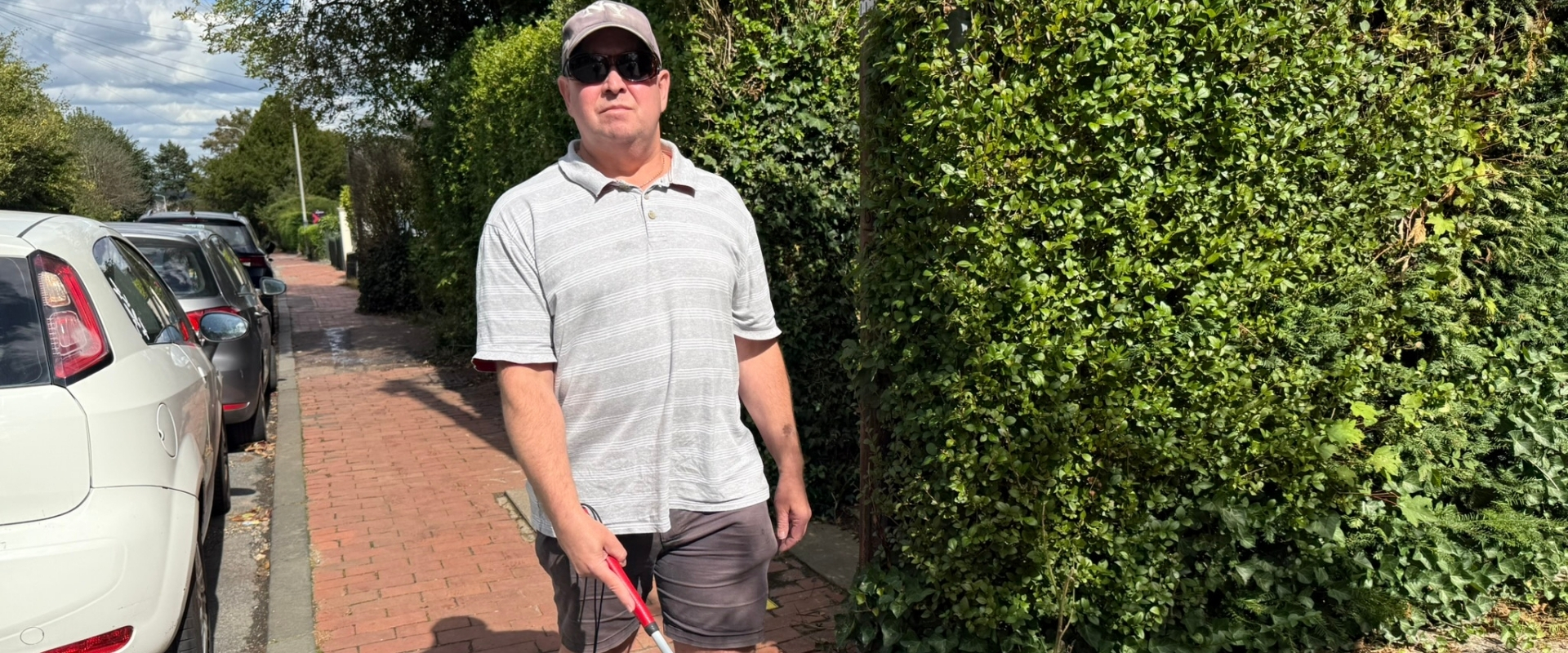 Tony walking along the path with his cane, whilst wearing his baseball cap and overspecs.