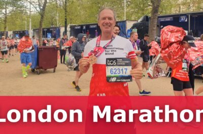 A KAB london marathon runner stands holding his medal and running bib, with lots of people walking behind him. Text reads: London Marathon