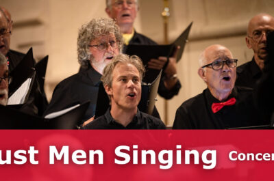 A group of men singing in a hall. They are holding choir books and are dressd smart with a black shirt and red bow tie.