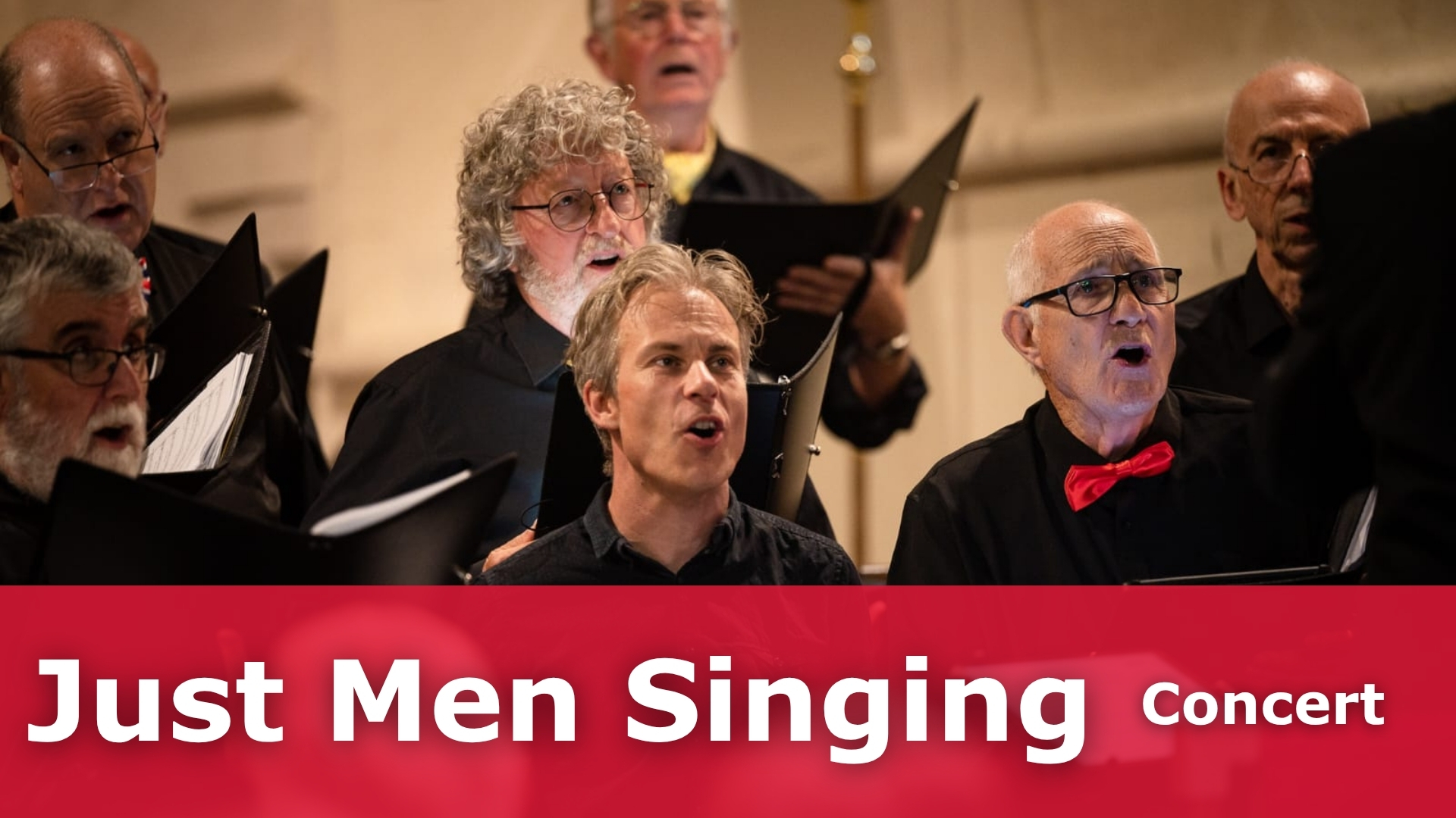 A group of men singing in a hall. They are holding choir books and are dressd smart with a black shirt and red bow tie.