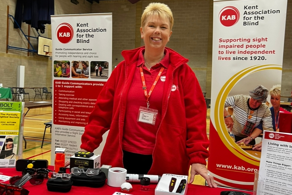 A KAB staff member standing behind a table at a previous exhibition. There are overspec glasses and magnifiers on the table in front of her.