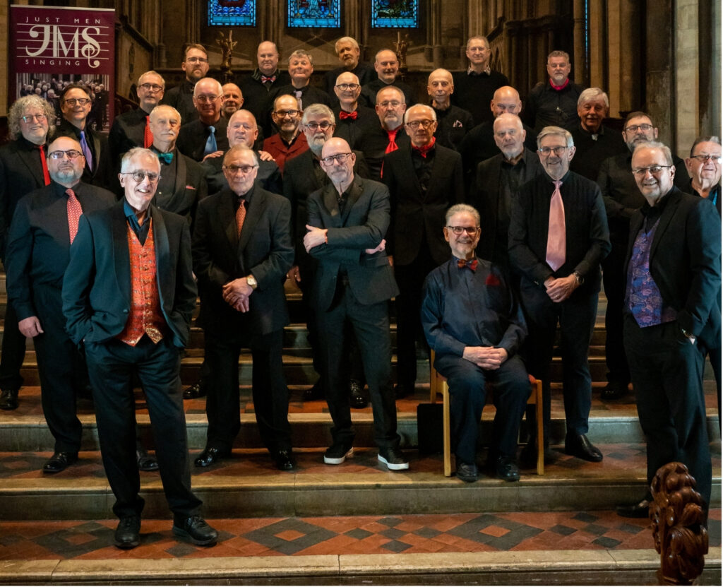 A big group of men wearing black suits in a church. They are the Just Men Singing Choir and they look happy.